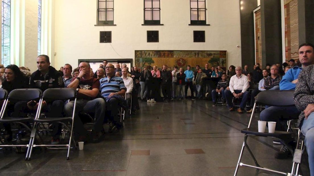Belangstellenden volgen de vergadering via een groot scherm