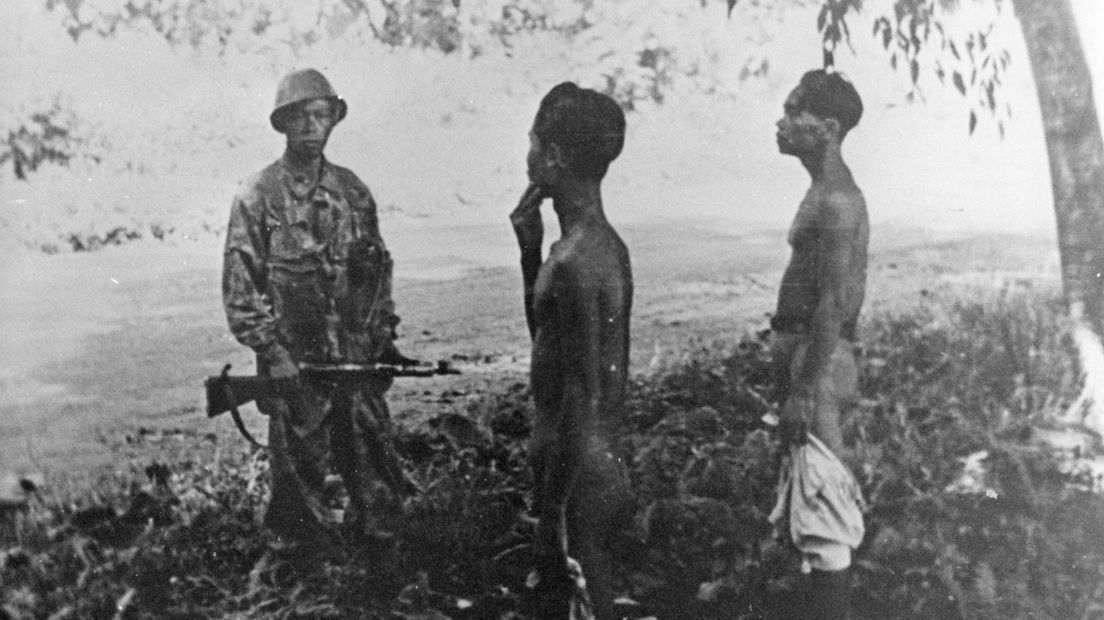 Een KNIL soldaat bewaakt twee gevangen die verhoord worden.