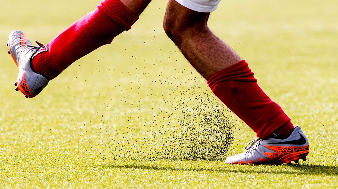 Een voetballer trapt de bal weg op een omstreden kunstgrasveld.
