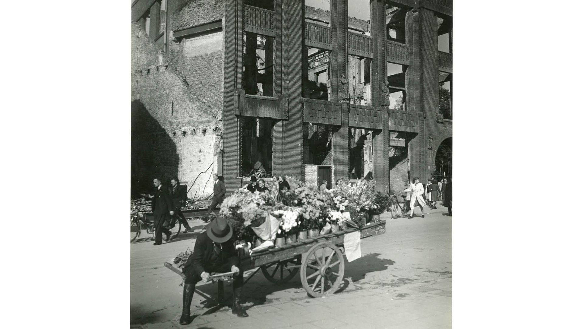 Een bloemenverkoper staat voor de uitgebrande gevel van de Levensverzekeringsmaatschappij in Arnhem, september 1945. (Gelders Archief: 4291-35, Emmy Andriesse, Public Domain Mark 1.0 licentie)