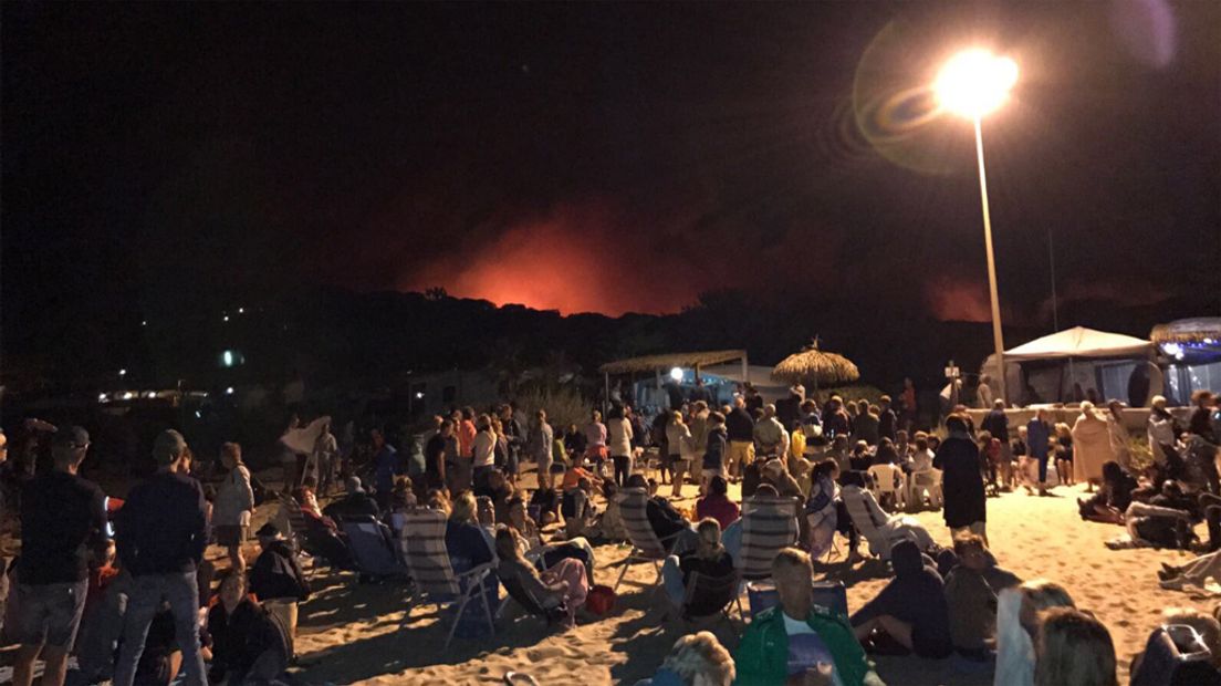 Alle campinggasten staan midden in de nacht op een strand te kijken naar de brand.
