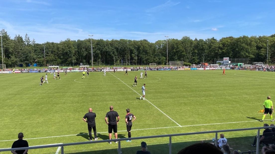 Toeschouwers en pers kijken geïnteresseerd toe vanaf de hoofdtribune op Sportpark Boerbos