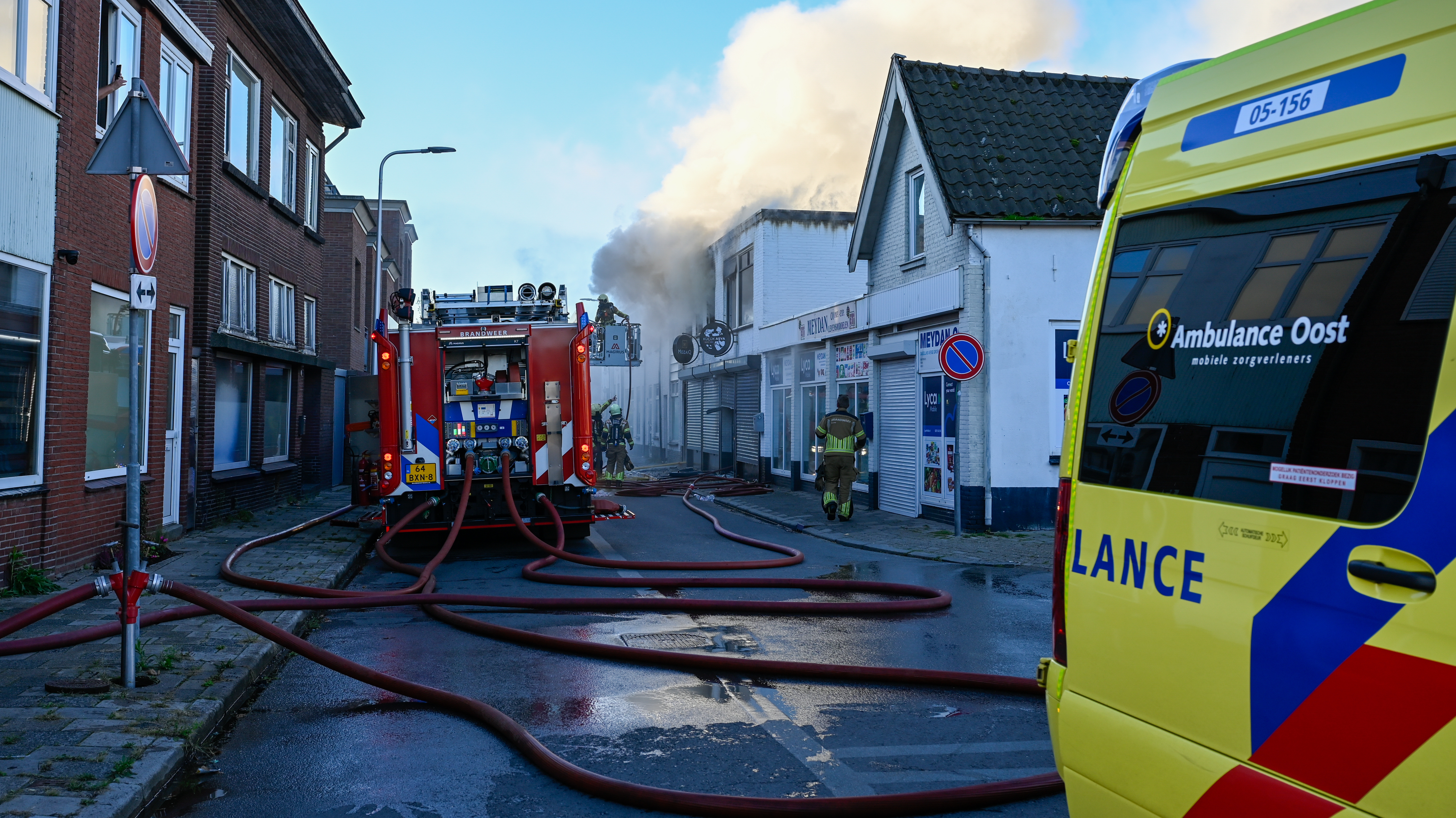 Terwijl de brandweer het vuur bestrijdt en dikke rookwolken opstijgen, staat een ambulance paraat op de Rohofstraat.