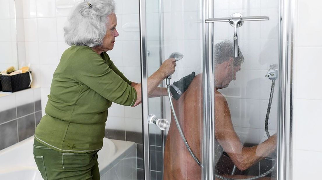 Een oudere man staat onder de douche en wordt gewassen door zijn vrouw.